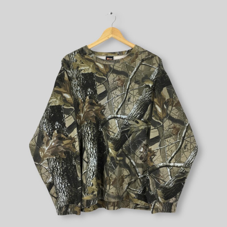 May include: A long-sleeved camouflage sweatshirt with a crew neck, hanging on a wooden hanger. The pattern features a mix of brown, green, and tan, resembling a forest scene. The sweatshirt is made of a soft material.