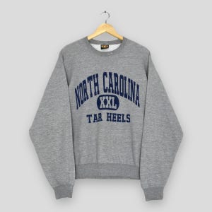 Puede incluir: Una sudadera gris con las palabras "NORTH CAROLINA" en azul marino arqueadas sobre el pecho, con "XXL" y "TAR HEELS" debajo. La sudadera est&aacute; colgada de una percha de madera.