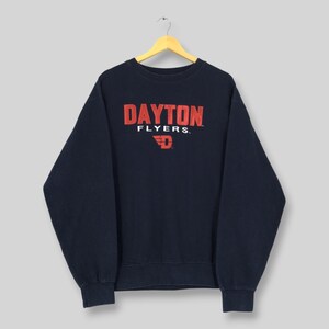 Könnte beinhalten: Marineblaues Sweatshirt mit dem Text "DAYTON FLYERS" in roten Blockbuchstaben. Darunter befindet sich ein rotes "D"-Logo. Das Sweatshirt hängt an einem Holzbügel.