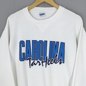 May include: White crew neck sweatshirt with the word "CAROLINA" in blue block letters and "Tar Heels" in a script font. The sweatshirt has a small "BIKE" tag near the neckline.