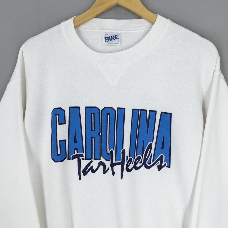 May include: White crew neck sweatshirt with the word "CAROLINA" in blue block letters and "Tar Heels" in a script font. The sweatshirt has a small "BIKE" tag near the neckline.