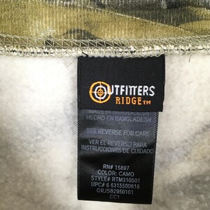 May include: A close-up of a tag from Outfitters Ridge clothing. The tag is black with white text and a logo. The tag reads "COLOR: CAMO" and "STYLE# RTM310501". The background is camouflage fabric.
