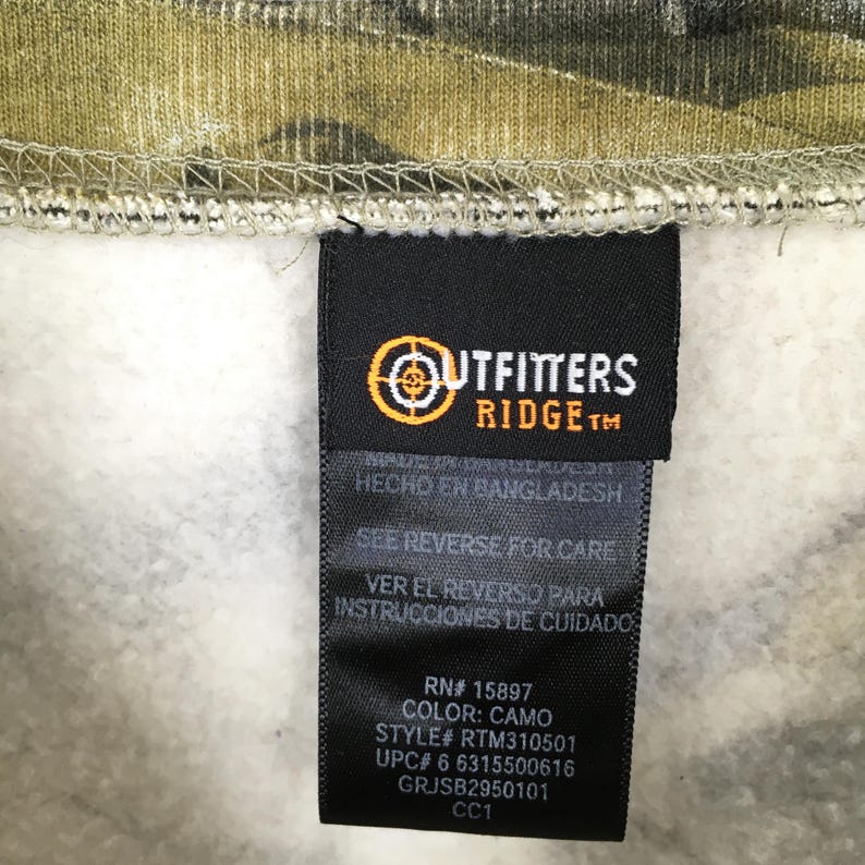 May include: A close-up of a tag from Outfitters Ridge clothing. The tag is black with white text and a logo. The tag reads "COLOR: CAMO" and "STYLE# RTM310501". The background is camouflage fabric.