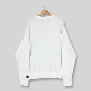 May include: A plain white long-sleeve crewneck sweatshirt hanging on a wooden hanger. The garment features a small, circular blue logo with white text on the lower left sleeve cuff. The sweatshirt has a simple, solid-coloured design throughout.