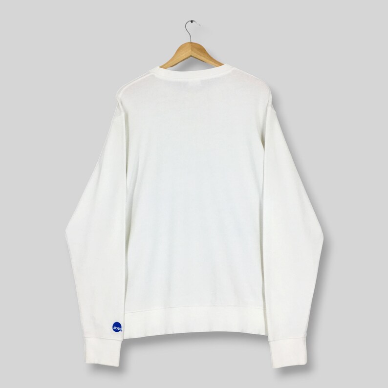 May include: A plain white long-sleeve crewneck sweatshirt hanging on a wooden hanger. The garment features a small, circular blue logo with white text on the lower left sleeve cuff. The sweatshirt has a simple, solid-coloured design throughout.