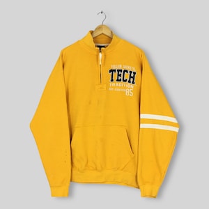 May include: Mustard yellow sweatshirt with a half-zip collar, a front pocket, and white stripes on the left sleeve. The sweatshirt features the text "YELLOW JACKETS TECH TRADITION EST. EIGHTEEN 85" in black and white.