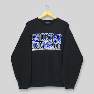 Puede incluir: Sudadera negra de cuello redondo con las palabras "SHORTER UNIVERSITY" en letras azules y blancas. Debajo del nombre de la universidad está el texto "TRANSFORMING LIVES THROUGH CHRIST". La sudadera está colgada en una percha de madera.