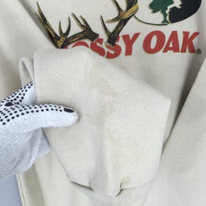 May include: Cream-coloured sweatshirt featuring the text "Mossy Oak" in red, along with a graphic of antlers and a tree. The sleeve is folded, showing a stain. The sweatshirt is made of a soft material.