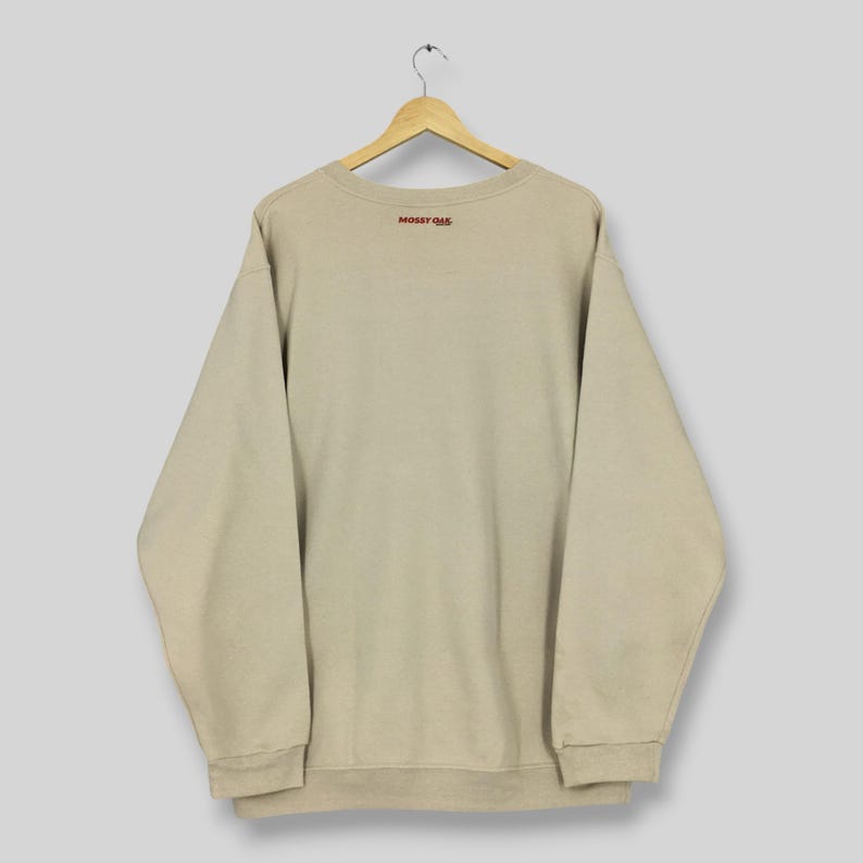 May include: A beige sweatshirt with the text "MOSSY OAK" in red lettering on the upper back. The sweatshirt is suspended from a wooden hanger against a light grey backdrop. The sweatshirt has long sleeves and a crew neck.