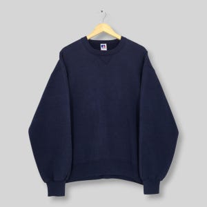 Könnte beinhalten: Ein marineblaues Sweatshirt mit Rundhalsausschnitt und langen Ärmeln, das an einem Holzbügel hängt. Das Sweatshirt hat ein kleines Logo auf der oberen linken Brust. Die Bündchen und der Saum sind gerippt.