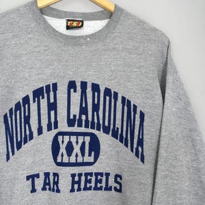 Puede incluir: Una sudadera gris con las palabras "NORTH CAROLINA" en azul marino arqueadas en el pecho, con "XXL" en un &oacute;valo azul marino y "TAR HEELS" debajo. La sudadera tiene cuello redondo y mangas largas.
