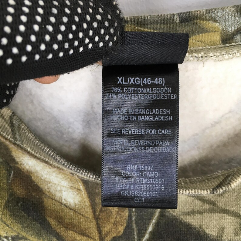 May include: Close-up of a tag on a camouflage-patterned sweatshirt. The tag is black with white text, including size XL/XG (46-48), fabric composition, and care instructions. The sweatshirt's pattern features shades of green, brown, and tan.