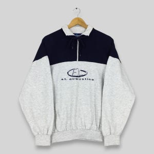 May include: A gray and navy blue sweatshirt with a half-zip collar. The sweatshirt features the embroidered text "FD st. augustine" in a stylized font. The sleeves and lower body are gray, while the upper chest and collar are navy blue.
