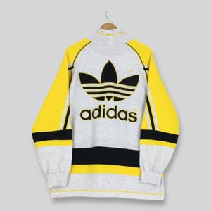 May include: A long-sleeve pullover featuring a heather gray body with bright yellow sleeves and shoulders. A large black Adidas trefoil logo and the word adidas are printed on the back. The garment includes black horizontal stripes on the sleeves.