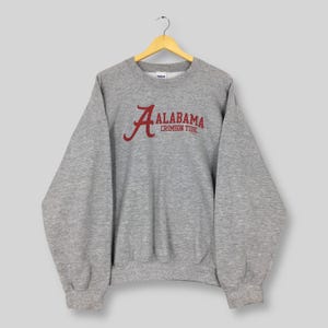 May include: A heather gray sweatshirt with the word "ALABAMA" in crimson red script, arched above "CRIMSON TIDE". The crew neck sweatshirt is hanging from a wooden hanger.