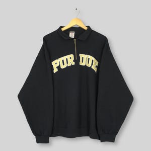 May include: Black sweatshirt with a collared neckline and a quarter-zip. The word "PURDUE" is printed across the chest in gold lettering. The sweatshirt is hanging on a wooden hanger.