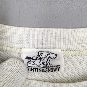 Peut inclure: Gros plan sur un sweat-shirt gris clair avec une &eacute;tiquette blanche repr&eacute;sentant une illustration en noir et blanc de Tintin et Milou. Le texte "TINTIN & SNOWY" est imprim&eacute; en dessous.