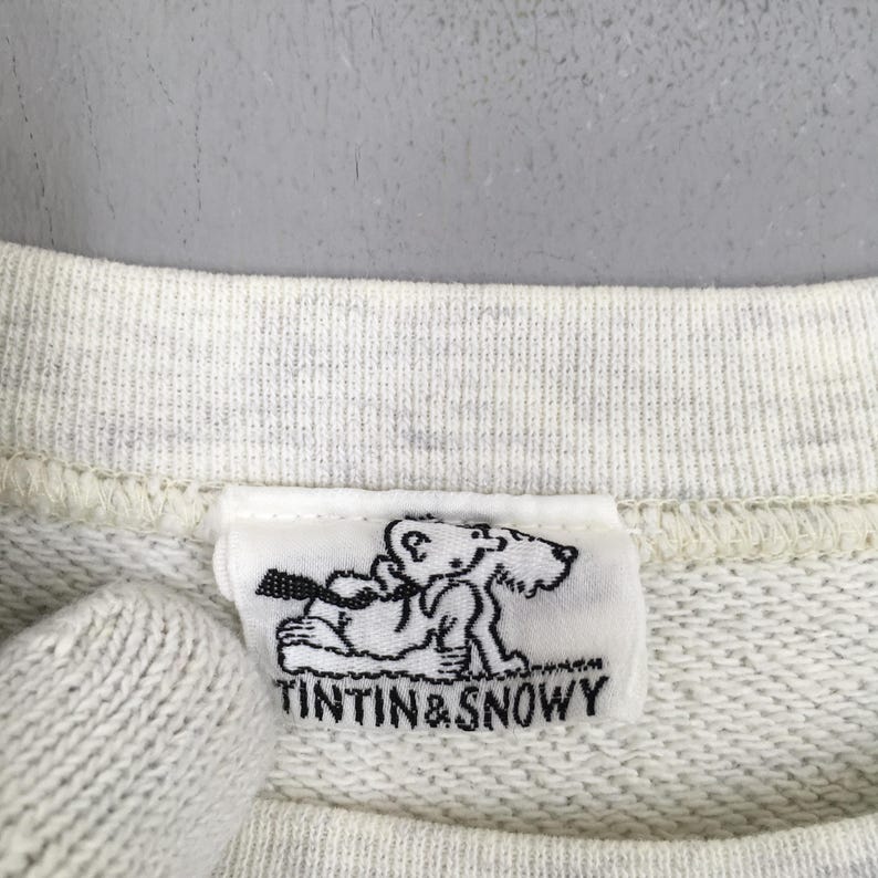 Peut inclure: Gros plan sur un sweat-shirt gris clair avec une &eacute;tiquette blanche repr&eacute;sentant une illustration en noir et blanc de Tintin et Milou. Le texte "TINTIN & SNOWY" est imprim&eacute; en dessous.
