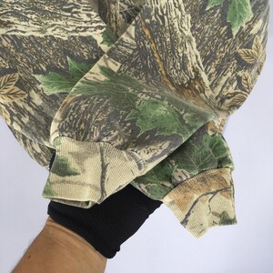 May include: Camouflage-patterned sweatshirt with shades of brown, green, and tan. The fabric features a realistic tree bark and leaf design. The sleeve cuff is tan and brown, with a black ribbed cuff underneath.