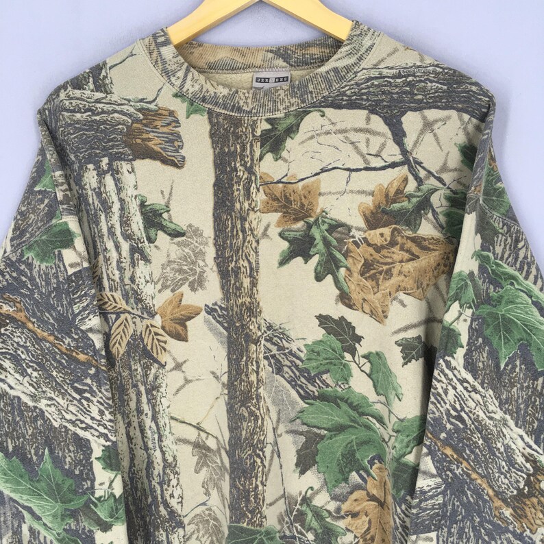May include: A tan and green camouflage sweatshirt with a crew neck. The pattern includes tree bark, leaves, and branches. The sweatshirt is made of a soft material and has a relaxed fit.