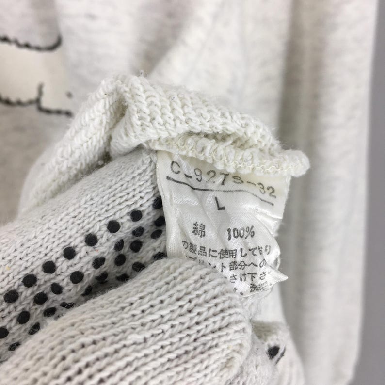 Peut inclure: Gros plan sur une &eacute;tiquette de v&ecirc;tement gris clair avec le texte "C-927S-32" et "L". L'&eacute;tiquette comprend &eacute;galement du texte japonais et les mots "綿 100%" indiquant 100% coton. Le v&ecirc;tement est gris clair.