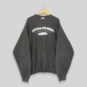 Puede incluir: Sudadera gris oscuro con el texto blanco "BRITISH COLUMBIA INSTITUTE OF TECHNOLOGY EST. 2004". La sudadera está colgada de una percha de madera sobre un fondo gris claro. Tiene mangas largas y cuello redondo.