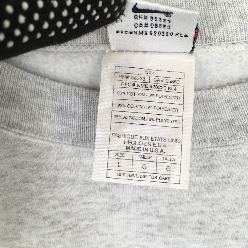 May include: Close-up of a grey sweatshirt label with the Nike logo. The label includes text with the RN, CA, and RFC numbers, along with the fabric composition: 95% cotton and 5% polyester. The label also states "Made in U.S.A."