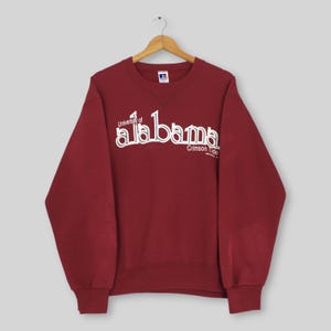 May include: A maroon long-sleeve crewneck sweatshirt featuring the white text University of Alabama Crimson Tide printed across the chest. The garment has a ribbed collar, cuffs, and hem, and is displayed on a wooden hanger against a plain background.
