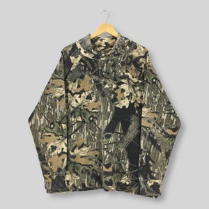 May include: A long-sleeved camouflage shirt with a mock neck collar. The shirt features a pattern of brown, green, and tan leaves and branches. The shirt is hanging on a wooden hanger against a light gray background.