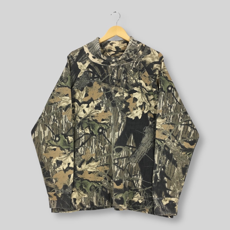 May include: A long-sleeved camouflage shirt with a mock neck collar. The shirt features a pattern of brown, green, and tan leaves and branches. The shirt is hanging on a wooden hanger against a light gray background.