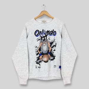 May include: A light gray sweatshirt featuring the word "Orlando" in blue and black lettering. The graphic depicts a cartoon wizard holding a basketball. The wizard is wearing a blue wristband and a black cape. The sweatshirt has long sleeves.