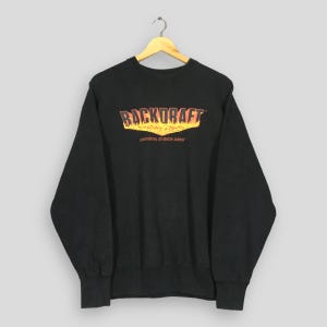 Peut inclure: Sweat-shirt noir à col rond avec l'inscription "BACKDRAFT" en orange et jaune flamboyant. En dessous, on peut lire "UNIVERSAL STUDIOS JAPAN" en blanc. Le sweat-shirt est suspendu à un cintre en bois.