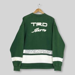 May include: A dark green long-sleeve sweatshirt featuring the white text TRD Sports on the upper back. A horizontal white band runs across the lower section with the word DEVELOPMENT printed in black letters inside a thin outlined border.