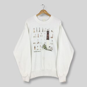 Può includere: Felpa bianca con una grafica colorata di vari fari. Il design include un faro marrone e bianco, circondato da illustrazioni di fari più piccoli. Il testo "Long Beach Island, NJ" è stampato sotto.