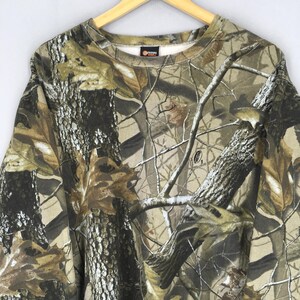 May include: A camouflage-patterned sweatshirt in shades of brown, green, and tan, featuring a crew neck and long sleeves. The design mimics a forest environment with tree bark and leaf details. The sweatshirt is hanging on a wooden hanger.