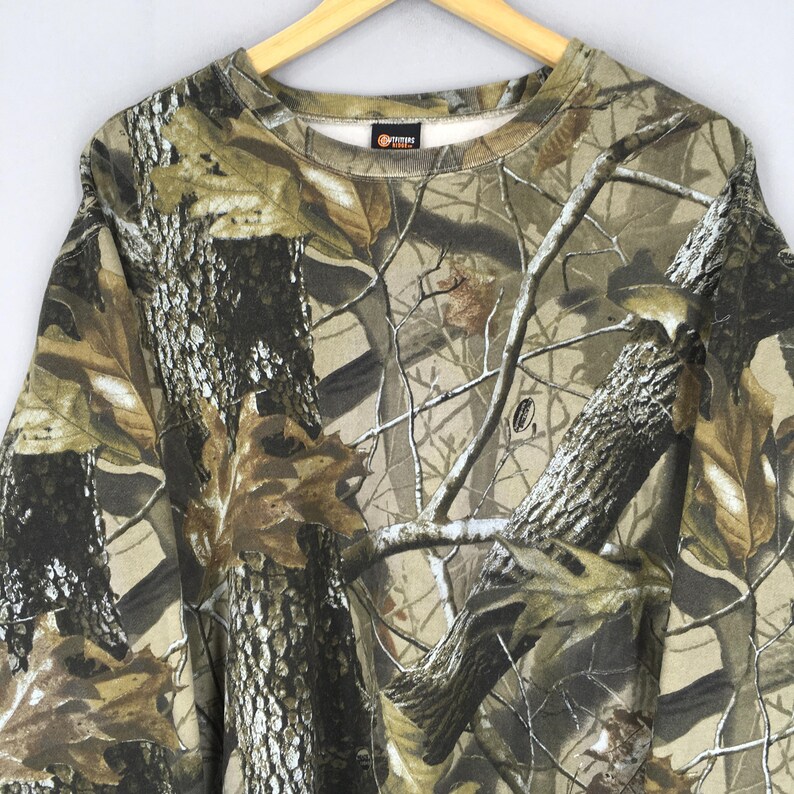 May include: A camouflage-patterned sweatshirt in shades of brown, green, and tan, featuring a crew neck and long sleeves. The design mimics a forest environment with tree bark and leaf details. The sweatshirt is hanging on a wooden hanger.