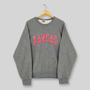 May include: A gray crewneck sweatshirt with the word "KANSAS" in red block letters with a white outline across the chest. The sweatshirt has long sleeves and ribbed cuffs and hem. The sweatshirt is hanging on a wooden hanger.