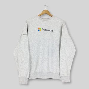 May include: A light gray heathered long-sleeve sweatshirt featuring the Microsoft logo in the center of the chest with the four-color square icon and the word Microsoft in dark gray text. A small #Microsoft hashtag is printed on the right sleeve.
