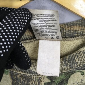 May include: Close-up of a tag on a camouflage-patterned sweatshirt. The tag includes washing instructions in multiple languages, including English, French, and Spanish. The sweatshirt is made of cotton and polyester.