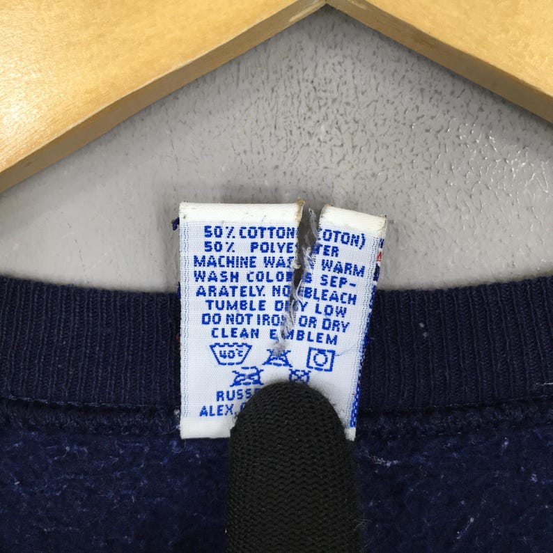 May include: Close-up of a navy blue garment with a white tag. The tag has text indicating the fabric composition, washing instructions, and the brand name "RUSS ALEX". The tag is torn in the middle.