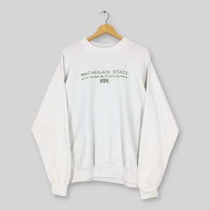 Puede incluir: Sudadera blanca de cuello redondo con el texto "MICHIGAN STATE SPARTANS STATE" en letras verdes. La sudadera está colgada de una percha de madera sobre un fondo gris claro. Tiene mangas largas y dobladillos acanalados.