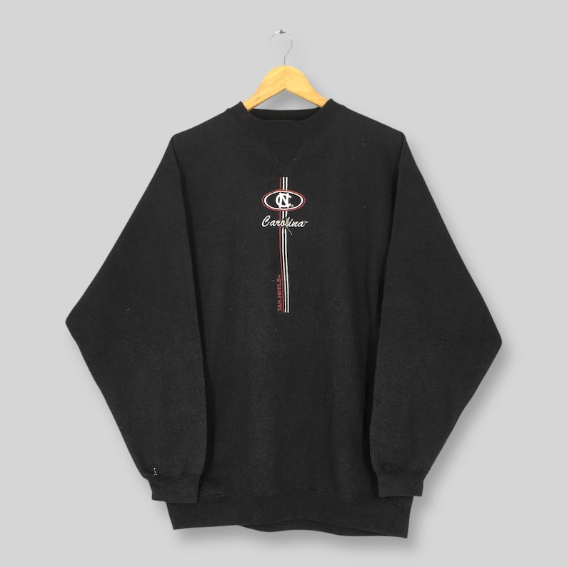 May include: Dark gray sweatshirt with the word "Carolina" in script and a logo featuring the letters "NC" in a circular design. The sweatshirt has a crew neck and long sleeves. The design includes red and white stripes.