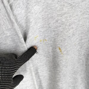 May include: Close-up of a pale grey sweatshirt with yellow stains. The sleeve and part of the shoulder are visible. A black glove with white polka dots is touching the fabric.