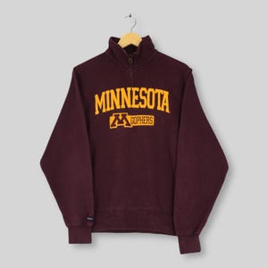 May include: Maroon quarter-zip sweatshirt with the text "MINNESOTA" in gold block letters and the "M Gophers" logo. The sweatshirt has a high collar and a zipper closure. The sleeves and hem have ribbed cuffs.