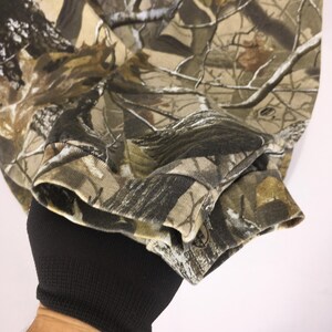 May include: Close-up of a long-sleeved camouflage shirt. The shirt features a detailed pattern of brown, green, and tan hues, mimicking a forest environment. The cuff of the sleeve is visible, with a ribbed texture and a small logo.