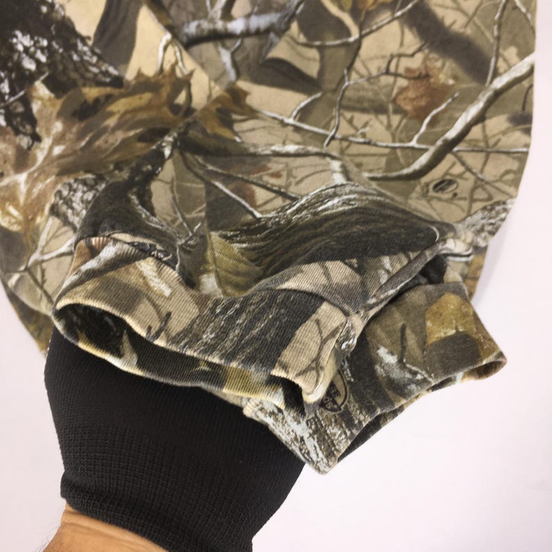 May include: Close-up of a long-sleeved camouflage shirt. The shirt features a detailed pattern of brown, green, and tan hues, mimicking a forest environment. The cuff of the sleeve is visible, with a ribbed texture and a small logo.