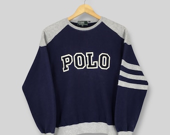 May include: Navy blue sweatshirt with gray raglan sleeves and trim. The word "POLO" is prominently displayed in white block letters across the chest. Three gray stripes accent one sleeve. A classic, vintage style.