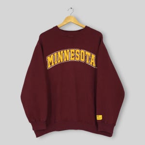 以下が含まれることがあります： 胸に「MINNESOTA」の文字が金色の文字でアーチ状に描かれた、マルーン色のスウェットシャツ。白い縁取りが施されています。スウェットシャツは長袖で、クルーネックです。右下に小さな黄色のタグが見えます。