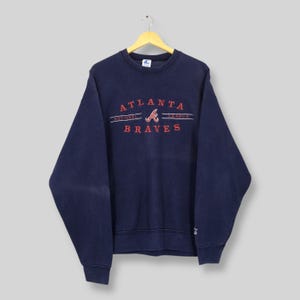 May include: A navy blue crew neck sweatshirt featuring the text "ATLANTA BRAVES" in red. The sweatshirt has a vintage aesthetic, with a slightly faded appearance. The team logo is also in red.