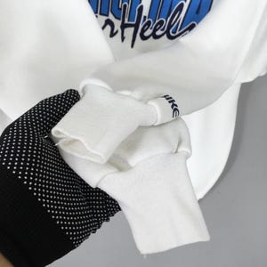 May include: White sweatshirt with blue text that reads "Heel". The sleeve cuff has the word "Nike" printed on it. The sweatshirt is being held by a hand wearing a black glove with white polka dots.